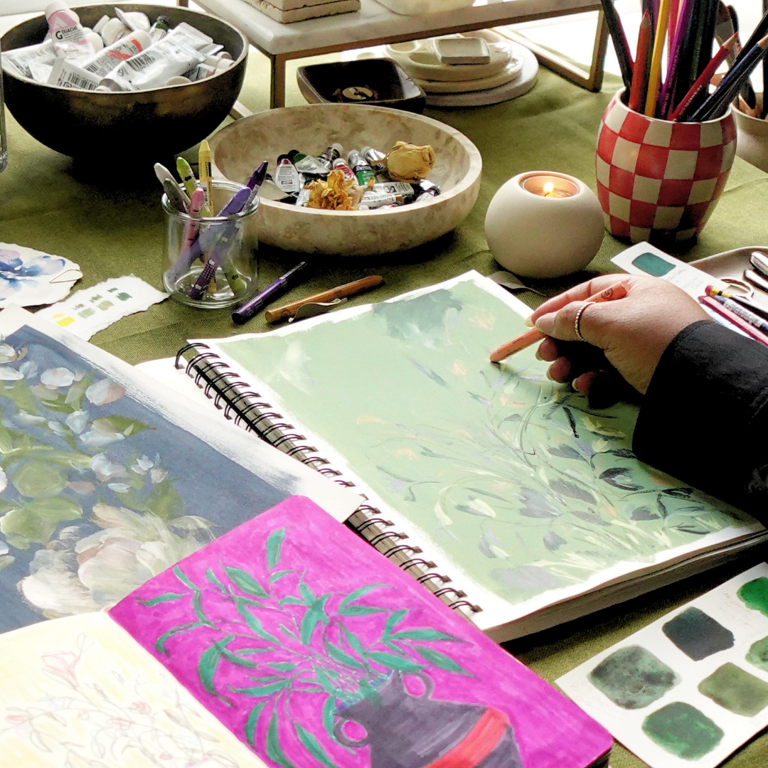 hand holding a pencil at desk with artwork of green leaves and a vase, alongside paints in marble bowls